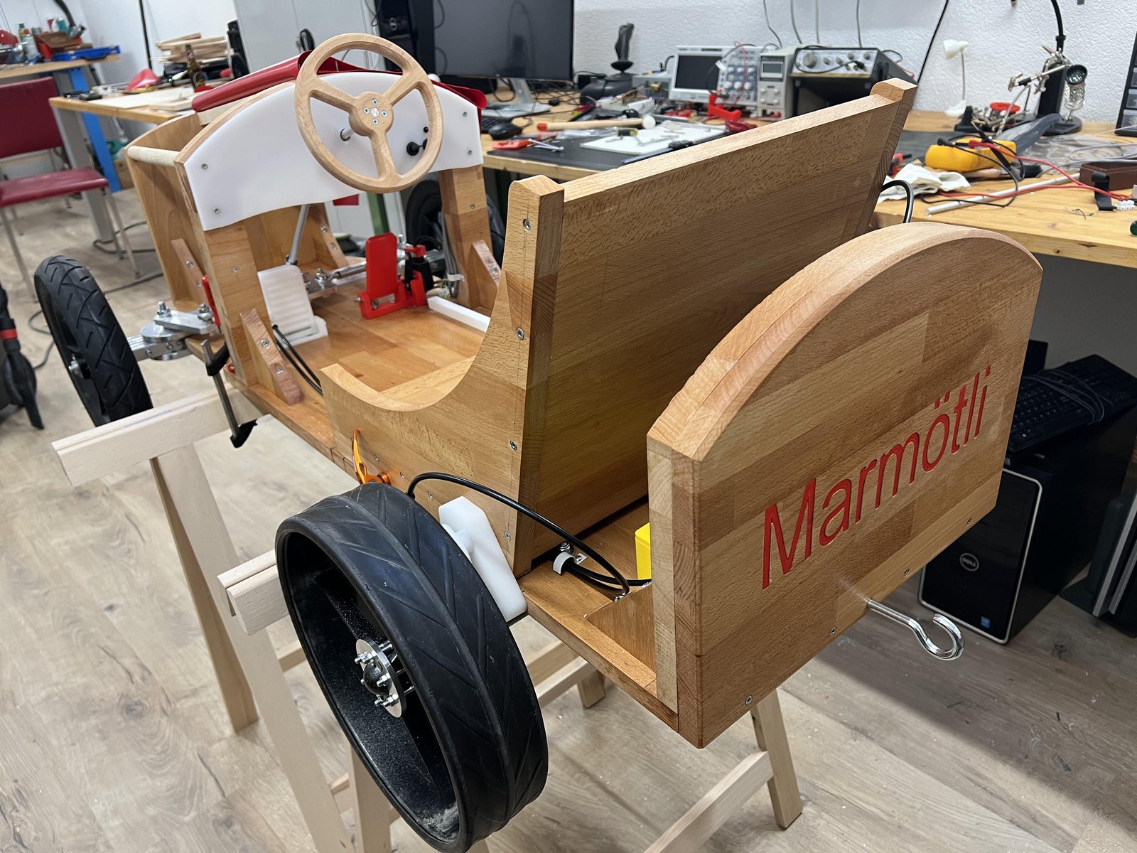 Marmötli im Rohbau: Holzkarosserie mit rotem Schriftzug, Lenkrad und Rädern auf Montageböcken
